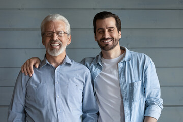 Fototapeta na wymiar Father and son portrait. Smiling mature dad young adult child hug look at camera. Two friendly relatives senior grandfather grown grandson posing for picture to family album on grey fence background