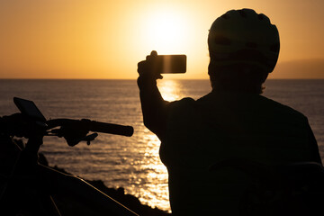Silhouette of man looking at sunset taking a photography with mobile phone, wearing sport helmet after cycling along the sea. © luciano