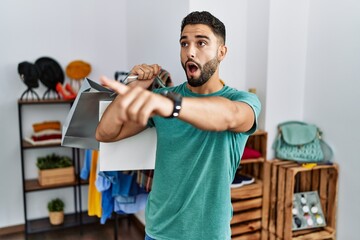 Young handsome man with beard holding shopping bags at retail shop pointing with finger surprised ahead, open mouth amazed expression, something on the front