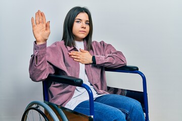Young brunette woman sitting on wheelchair swearing with hand on chest and open palm, making a...