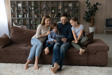 Happy family with children using tablet together, relaxing on cozy couch at home, smiling parents with daughter and son spending leisure time holding device, having fun online, looking at screen