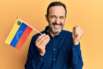 Middle age hispanic man holding venezuelan flag screaming proud, celebrating victory and success very excited with raised arm
