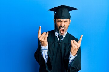 Middle age hispanic man wearing graduation cap and ceremony robe shouting with crazy expression doing rock symbol with hands up. music star. heavy concept.