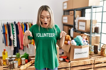 Asian young woman wearing volunteer t shirt at donations stand pointing down with fingers showing advertisement, surprised face and open mouth