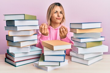 Young caucasian woman sitting on the table with books doing money gesture with hands, asking for...