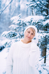 Fototapeta na wymiar Outdoor winter portrait of happy smiling woman posing in park in front of fir tree. Model wearing warm white sweater.