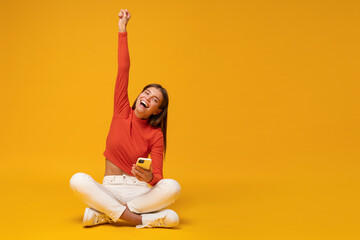 Winner. Excited happy woman sitting on floor with phone, shouting yes, isolated on yellow