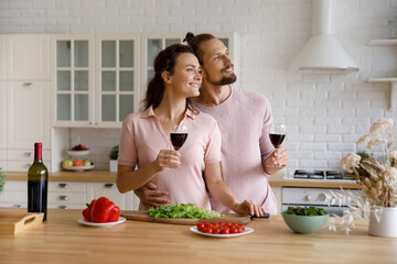 Loving bonding affectionate millennial married family couple holding wineglasses in hands, looking in distance imagining planning visualizing future, enjoying carefree home date in modern kitchen.