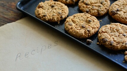 Crispy freshly baked oatmeal cookies with pieces of chocolate on a baking sheet on a table with a sheet of yellowed paper with the inscription Recipes.