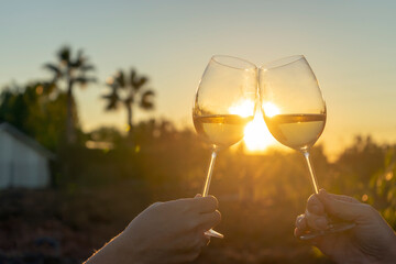 TWO HANDS HOLDING WINE GLASSES AND TOASTING. SUNSET WITH PALM TREES IN THE BACKGROUND. LIFESTYLE CONCEPT.
