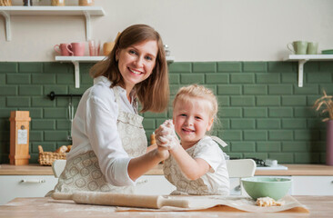 A young mother and her little daughter are cooking together in the kitchen. Mom and child prepare baking dough at home