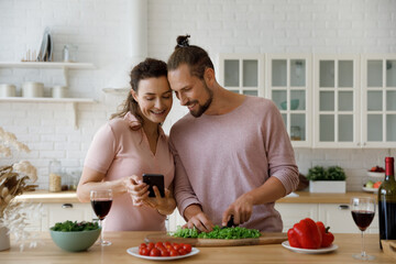 Happy young beautiful woman holding cellphone in hands, showing video with food recipe or sharing funny photo content with laughing husband, enjoying cooking together in modern kitchen on weekend.