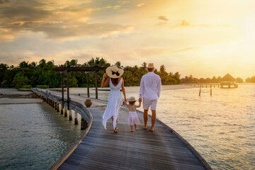 A family in white summer clothes walks holding hands over a wooden pier towards a tropical island...