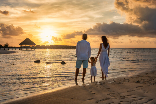 A family in white summer clothing stands hand in hand on a tropical paradise beach during sunset time and enjoys their vacation time in the Maldives islands
