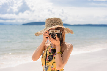 Short-haired Asian woman in a yellow floral dress and straw hat standing on the beach using camera take a photo in front of her with a blurry sea, mountain and cloudy sky background on a sunny day.