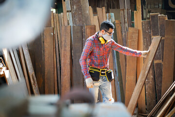Yong Caucasian male carpenter in safety goggles, face mask, and ear muffs around his neck standing holding a wooden plank with looking, checking surface and quality of a wooden plank in a wood factory - Powered by Adobe
