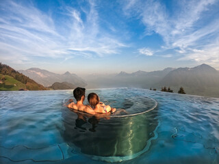 Back of young romantic Asian couple sitting together on an underwater seat in the swimming pool admired beautiful seascapes surrounded by mountains with amazing blue sky and soft clouds on a clear day