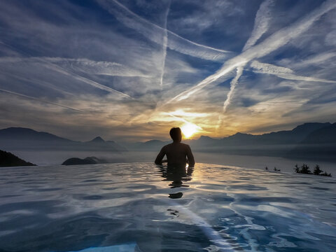 Back of young Asian man without shirt standing in the modern swimming pool admired calm beautiful seascapes surrounded by mountains with amazing cloud appeared as a line in the blue sky during sunset - Powered by Adobe