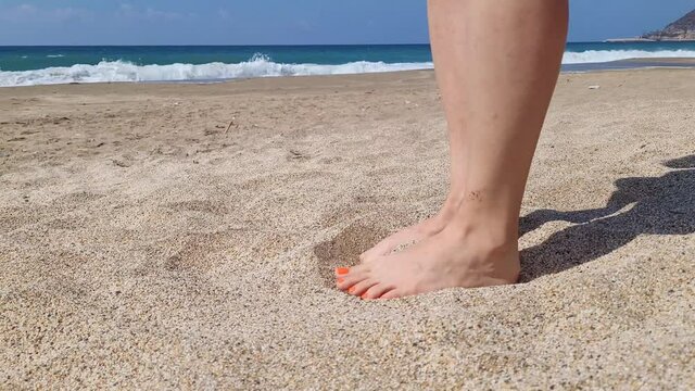 Women's feet are on the beach in the sand, the sea in the background.