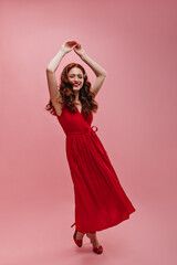 Full-length portrait of young european woman actively moving against pink background. Flawless beauty with dark wavy hair is wearing long red dress and shoes. 