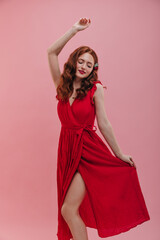 Pretty young european woman stands on pink background with her hand up. Red-haired lady, looking down, holds skirt from red dress, exposing her light-skinned thin leg. Good mood, fashion trends