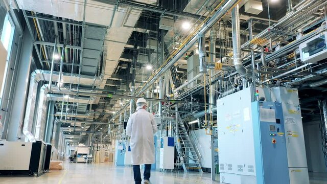 Male specialist in a lab coat is observing factory machines and pipes