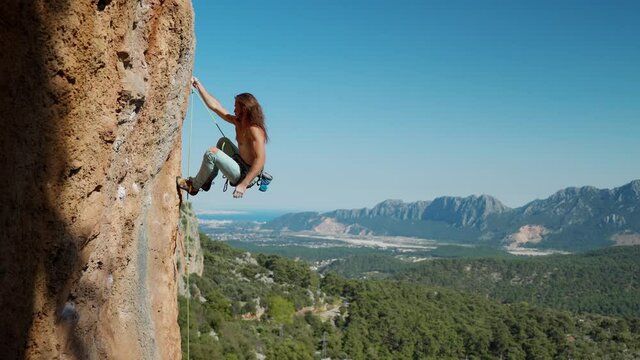 attractive strong and fit man rock climber with long hair hangs on rope on vertical cliff and looks up. beautiful landscape with rocky mountain ridge and clear blue sky on background