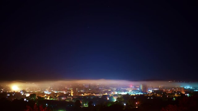  Fog on the night city and stars in the sky, loop video. Time lapse of fog and a starry sky in motion above the lights of a city skyline on european city