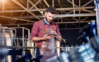 Young male brewer in leather apron supervising the process of beer fermentation at modern brewery factory