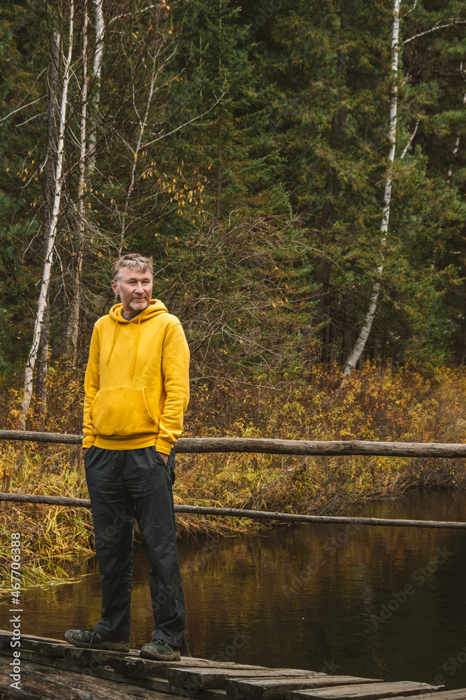 Wall mural A middle-aged traveler in yellow hoodie stands on wooden bridge over a river in the forest, enjoying beautiful autumn day outdoors. Local travel and weekend hikes. Sustainable environment. Vertical. - Wall murals