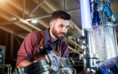 Young male brewer in leather apron supervising the process of beer fermentation at modern brewery factory