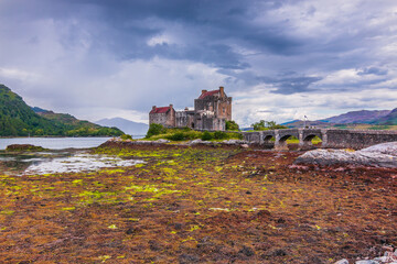 Naklejka na ściany i meble Lake Loch Duich at low tide. Eilean Donan Castle in summer on the island. Castle and a stone bridge and small archways. Algae on stones in the foreground and hills in the background with clouds