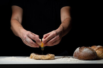 Cooking the bread. The chef's hands add a chicken egg that flies onto the white table with the flour and dough. Chef sprinkles white flour dust with a man's hand on a black background. Space for text.