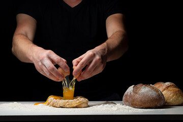 Cooking the bread. The chef's hands add a chicken egg that flies onto the white table with the flour and dough. Chef sprinkles white flour dust with a man's hand on a black background. Space for text.