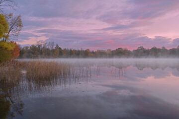 Fototapeta premium Foggy autumn landscape at dawn of the shoreline of Whitford Lake with mirrored reflections in calm water, Fort Custer State Park, Michigan, USA