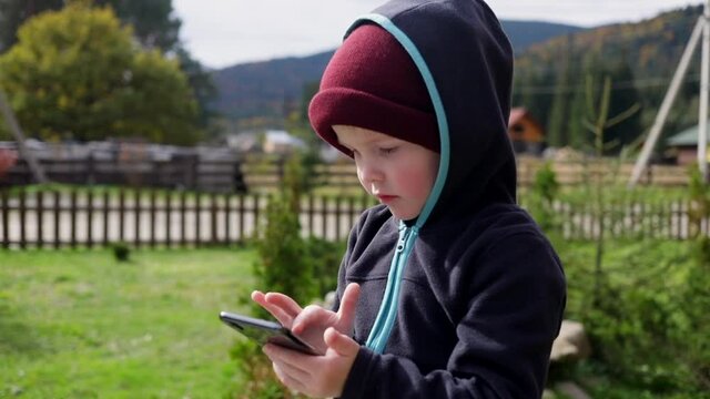 Little blonde boy using smartphone mobile looking photos tapping touchscreen. Closeup portrait. Beautiful mountains on sunny background. Recreation activity. Traveling family. Internet connect.
