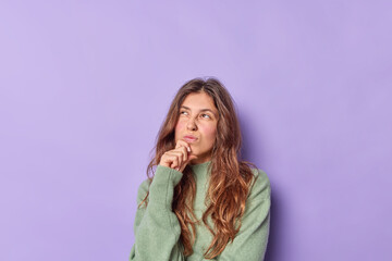 Thoughtful young Caucasian woman keeps hand on chin frowns face thinks above something concentrated above wears casual jumper isolated over purple background copy space for your advertising content