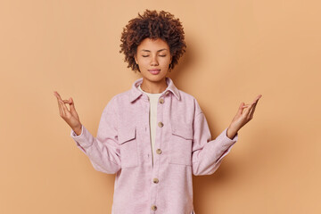 Calm relaxed curly haired woman stands with closed eyes meditates and enjoys peaceful atmosphere makes mudra gesture wears pink jacket isolated over beige background. Harmony in life and balance