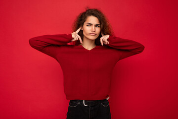 Photo of young dissatisfied tired attractive brunette wavy-haired woman with sincere emotions wearing casual red sweater isolated on red background with empty space and covering ears
