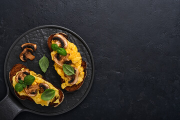 Scrambled eggs with fried mushrooms and basil on bread on black table background. Homemade breakfast or brunch meal - scrambled eggs and mushrooms sandwiches. Top view with copy space
