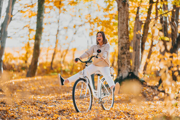Young pretty woman riding retro-styled white bicycle in golden autumn park. Amazing funny scene of...
