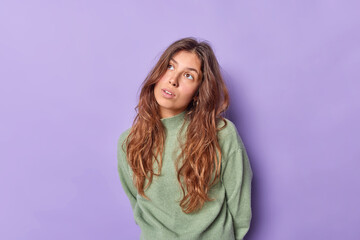 Pretty brunette young European woman concentrated above with thoughtful expression poses against purple background recalls something in mind tries to remember her childhod. People and thoughts