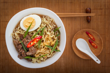 Ramen soup with chicken, noodles, egg, hot pepper and sprouted beans in a white plate against a mat with a spoon and chopsticks. Asian street food. View from above