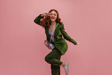 Happy caucasian woman showing victory sign near face smiling strongly on pink background. Long-haired red-haired beauty laughs with her leg curled up at knee, dressed in jacket, pants and top.