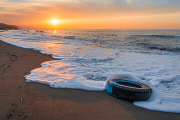 Fototapeta na wymiar A rubber car wheel on the beach among the waves, sunset from the Mediterranean sea coast, the sea polluted with garbage, sandy beach and orange sky and clouds, landscape.