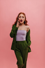 Modern caucasian young lady is shocked by what she saw, opening her mouth on pink background. Red-haired girl in lilac top and dark green fashionable suit. Emergence of positive emotion, concept