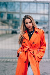 Fashionable blonde woman wearing orange coat, blue jeanse , white sneackers and neckchain posing in street