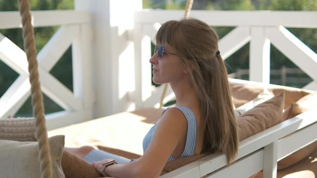 Young happy brunette woman lying down on terrace sofa with soft pillows enjoying warm summer day. Concept of free time on fresh air