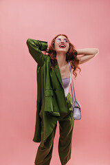 Happy pretty caucasian young woman holding hands behind her head smiling broadly with teeth on pink background. Beauty raised her head up, closing eyes, wearing top, jacket on one shoulder and pants.