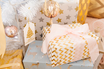 Heap of stack of gift boxes, wrapped in gift paper, stands under white Christmas tree with decorations. Festive decor, Christmas concept.